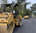 Steamroller on a newly paved street, cars and workers in the background working on repaving the street