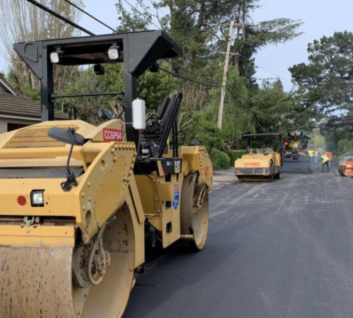 Steamroller on a newly paved street, cars and workers in the background working on repaving the street